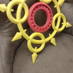 May include: A yellow and red 3D printed design with a circular shape and pointed edges. The design is reminiscent of a sun or a star.