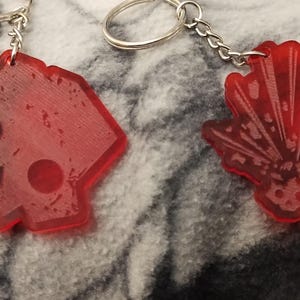May include: Two red acrylic keychains. One keychain is shaped like a geometric figure with two black circles. The other keychain is shaped like a stylized explosion with a red and white pattern.