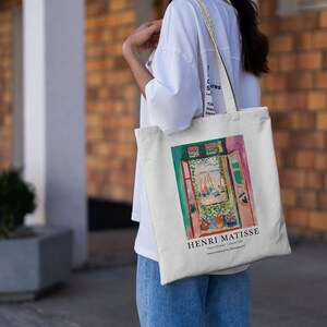 May include: A white canvas tote bag with a colorful painting of an open window with a view of boats on the water. The text "HENRI MATISSE" and "Open Window, Collioure 1905" is printed on the bag.