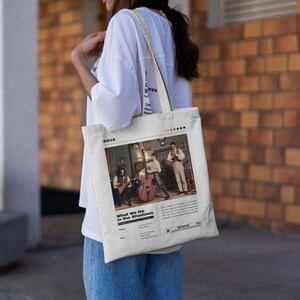 May include: A white tote bag with a band's album cover printed on it. The album cover features a band of three musicians playing instruments in a dimly lit room. The album title is "What We Do in the Shadows" and the year "2014" is printed at the top.