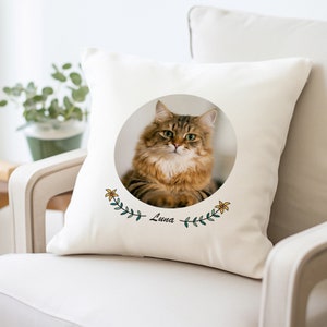 May include: A white pillow with a circular image of a brown tabby cat with green eyes. The cat is surrounded by a green wreath with yellow flowers and the name "Luna" is written below the wreath.