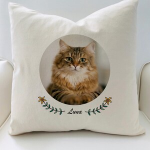 May include: A white pillow with a round image of a brown tabby cat with green eyes. The image is surrounded by a green leafy garland with yellow flowers and the name "Luna" written below.
