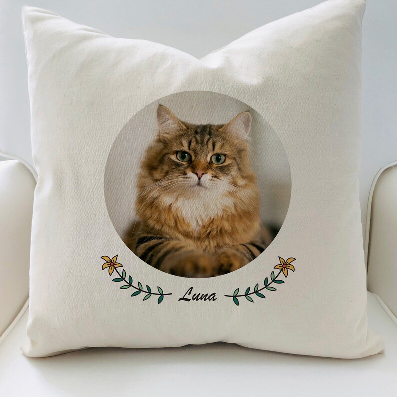 May include: A white pillow with a round image of a brown tabby cat with green eyes. The image is surrounded by a green leafy garland with yellow flowers and the name "Luna" written below.