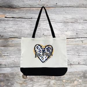May include: A white tote bag with black handles and a black bottom. The bag features a heart-shaped design with a leopard print pattern in blue and yellow, and the words "Be Kind" in black script.