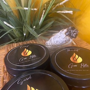 Op de afbeelding: Drie zwarte potten met body butter met gouden etiketten. De etiketten hebben een vlamontwerp en de tekst "Game Butter" en "Ultra Hydrating Body Butter". De potten zijn gelabeld met "Courage", "Manifest" en "Ground" met de tekst in wit.