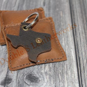 May include: A leather keychain in the shape of Texas, with a silver keyring, rests on a small, brown leather pouch. The keychain is dark brown and has a metal rivet. The pouch is a lighter shade of brown and has stitched edges. The background is a wooden surface.