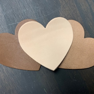 May include: Three heart-shaped leather pieces in different shades of brown. The largest heart is a light beige color, while the two smaller hearts are a darker brown.