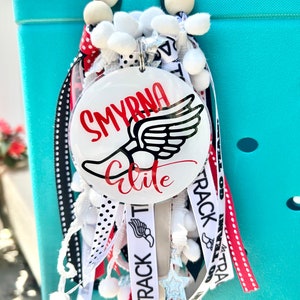 May include: White, black, and red ribbon charm with a white circle that says "SMYRNA elite" in black and red lettering with a black and white wing graphic. The charm is attached to a white tote bag.