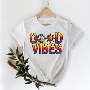 Puede incluir: Camiseta blanca con un diseño degradado arcoíris que dice "GOOD VIBES" con un símbolo de paz, un corazón y un sol.