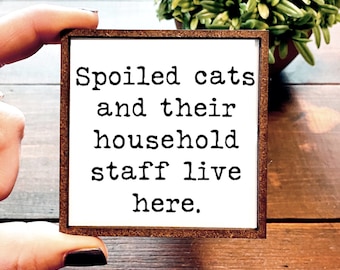 May include: A white wooden sign with brown trim reads "Spoiled cats and their household staff live here."