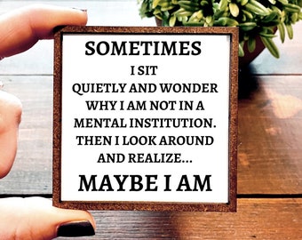 May include: A white wooden sign with brown trim, featuring the text "SOMETIMES I SIT QUIETLY AND WONDER WHY I AM NOT IN A MENTAL INSTITUTION. THEN I LOOK AROUND AND REALIZE... MAYBE I AM"