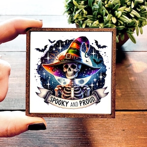 May include: A square wooden sign featuring a skeleton wearing a colorful witch hat, set against a starry night sky with bats. The banner reads "Spooky and Proud."