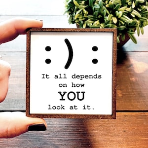 May include: A white wooden sign with a black and white smiley face and the text "It all depends on how YOU look at it."