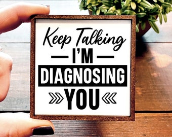 May include: A white wooden sign with a brown frame. The sign says "Keep Talking - I'm Diagnosing You" in black text.