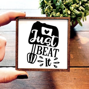 May include: A white wooden sign with a black silhouette of a stand mixer and the text "Just Beat It" in a black cursive font.