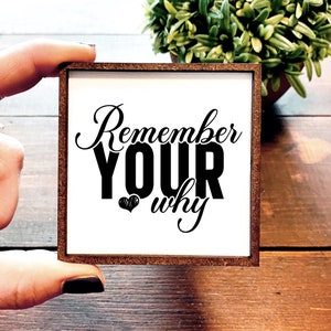 May include: A white wooden sign with a brown frame. The sign has black text that reads "Remember your why" with a small heart symbol.