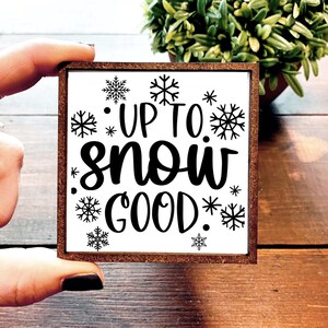 May include: A white wooden sign with a brown frame. The sign says "Up to snow good" in black cursive lettering. The sign is decorated with black snowflake designs.
