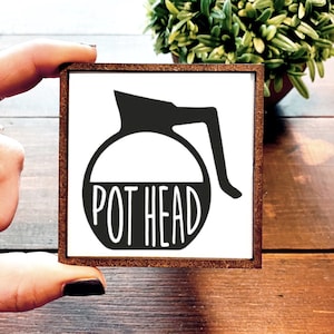 May include: A white square sign with a black silhouette of a coffee pot and the text "POT HEAD" in a white outline.