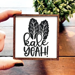 May include: A white wooden sign with a brown frame features a black silhouette of two kale leaves and the text "kale yeah!"