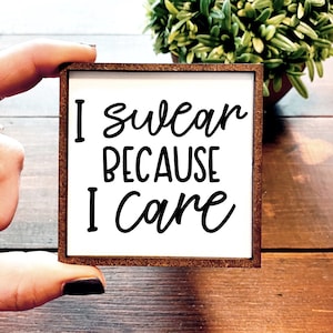 May include: A white wooden sign with a brown frame reads "I swear because I care" in black cursive font.