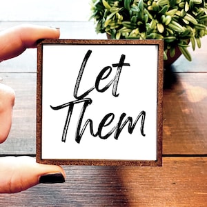 May include: A white wooden sign with a brown frame. The sign has black calligraphy that reads "Let Them".
