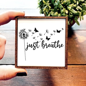 May include: A white wooden sign with a black silhouette of a dandelion and butterflies with the text "just breathe".