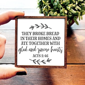 May include: A white wooden sign with a brown frame features a black and white quote from the Bible, Acts 2:46, "They broke bread in their homes and ate together with glad and sincere hearts."