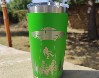 May include: A green stainless steel tumbler with a laser-etched design of a Bigfoot being abducted by a UFO. The design also includes a forest scene.