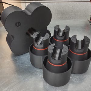 May include: A set of black 3D-printed components. The set includes a large, four-lobed piece and four smaller cylindrical pieces with a U-shaped top and a red accent ring. The items are arranged on a metallic surface.