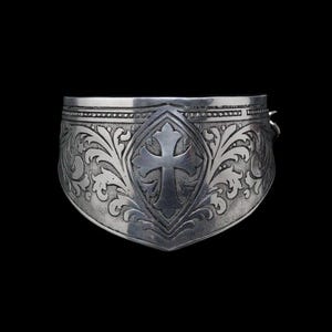 Peut inclure: Un bracelet manchette argenté avec un motif de croix gravée au centre. Le bracelet présente des motifs floraux complexes et une forme de bouclier, sur fond noir.