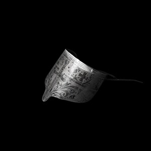 May include: A detailed, grayscale image of a decorative metal object, possibly a helmet or headpiece. The object features intricate floral and geometric patterns. The design includes a pointed front and a curved, raised section. The background is solid black.