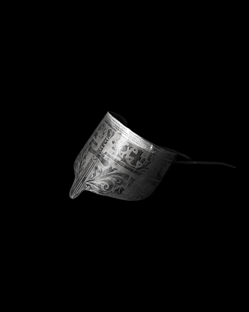 May include: A detailed, grayscale image of a decorative metal object, possibly a helmet or headpiece. The object features intricate floral and geometric patterns. The design includes a pointed front and a curved, raised section. The background is solid black.