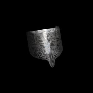 May include: A silver-colored, ornate metal object with intricate floral and geometric patterns. The object has a curved shape with a pointed end and appears to be a decorative piece or a part of a larger item. The surface reflects light, highlighting the detailed engravings.