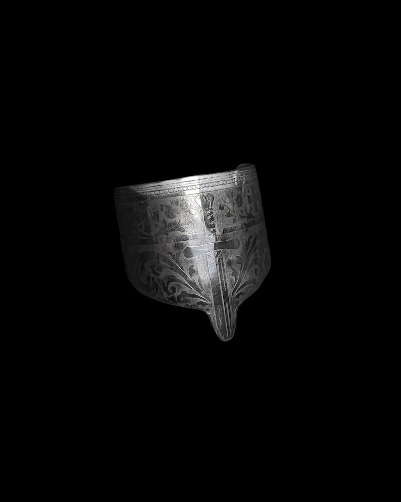 May include: A silver-colored, ornate metal object with intricate floral and geometric patterns. The object has a curved shape with a pointed end and appears to be a decorative piece or a part of a larger item. The surface reflects light, highlighting the detailed engravings.