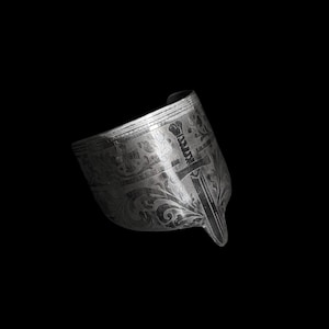 May include: A silver-toned cuff bracelet with intricate engraved floral and cross patterns. The cuff has a wide band with a pointed end, and the design is detailed with dark accents, creating a vintage aesthetic.