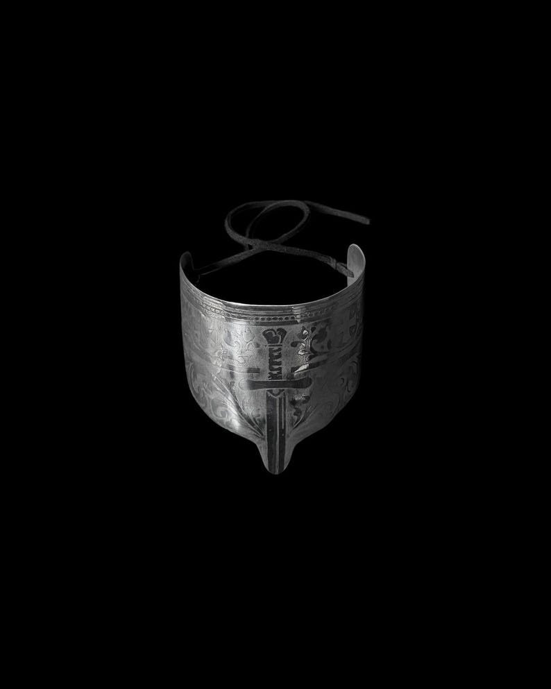 May include: A silver-toned metal cuff bracelet with intricate engraved patterns and a sword design. The bracelet has a curved shape and a black cord for closure, set against a black background.