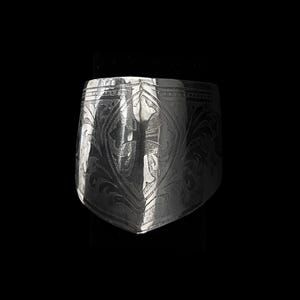 Peut inclure: Une bague argentée avec un motif floral gravé. La bague a une forme de bouclier avec un fond pointu et une large bande. Le design complexe recouvre toute la surface de la bague.