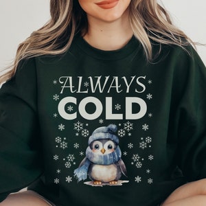May include: A dark green sweatshirt with a white penguin graphic and the text "*ALWAYS* COLD*" surrounded by snowflakes.