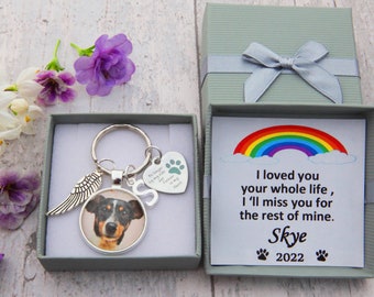 May include: A silver keychain with a paw print charm, a small silver charm with the letter "S", a silver angel wing charm, and a round photo of a dog. The keychain is in a small gray box. The box is next to a larger gray box with a white card inside. The card has a rainbow and the text "I loved you your whole life, I'll miss you for the rest of mine. Skye 2022".