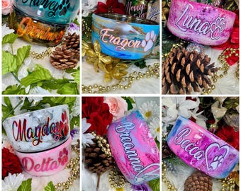 May include: A collection of personalized pet bowls featuring various colorful marbled and glittery finishes. Each bowl is customized with a name in script lettering and a paw print or heart icon, displayed among decorative pinecones and flowers.