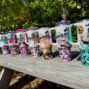 May include: A row of seven white insulated tumblers with handles and lids, each featuring a unique colorful marbled pattern, a leopard print base, a sunflower graphic, and the word Bronco written in a cursive black script font on the side.