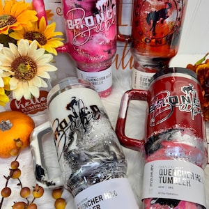 May include: Several Stanley Quencher H2.0 tumblers with handles are displayed. The tumblers feature various designs, including pink and black marble, silver glitter, and the text "Bronco Babe." One tumbler has a red and black design with the Ford Bronco logo.