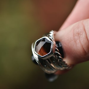 May include: A silver ring with a hexagonal brown gemstone set in the center. The ring is stacked with other silver rings.