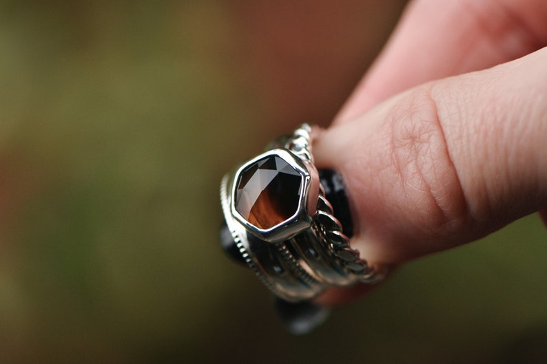 May include: A silver ring with a hexagonal brown gemstone set in the center. The ring is stacked with other silver rings.