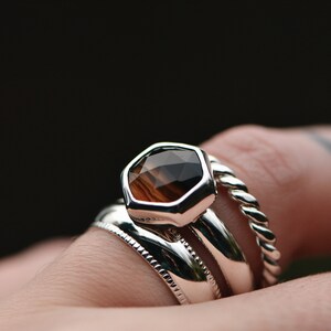 May include: A silver ring with a hexagonal brown gemstone set in the center. The ring is worn on a finger with a tattoo. The ring is stacked with two other silver bands.
