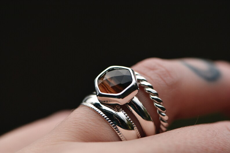 May include: A silver ring with a hexagonal brown gemstone set in the center. The ring is worn on a finger with a tattoo. The ring is stacked with two other silver bands.