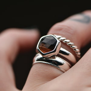 May include: A silver ring with a hexagonal brown gemstone set in a bezel setting. The ring is worn on a finger with a tattoo. The ring is stacked with two other silver bands.