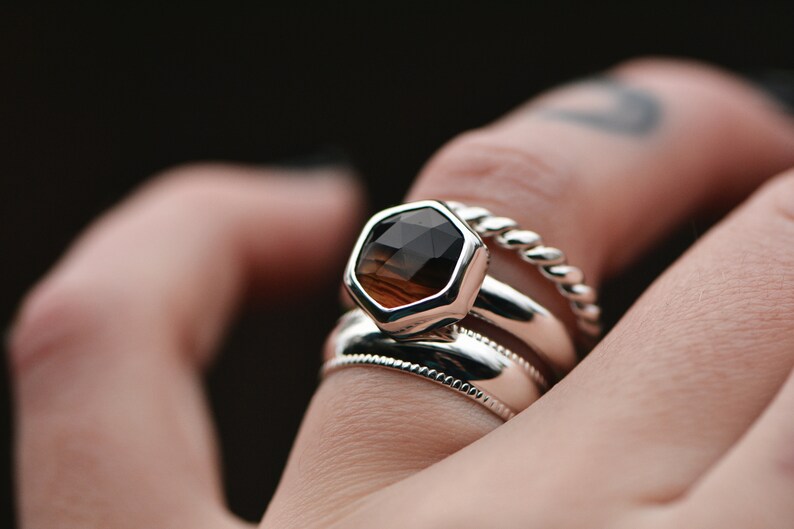 May include: A silver ring with a hexagonal brown gemstone set in a bezel setting. The ring is worn on a finger with a tattoo. The ring is stacked with two other silver bands.