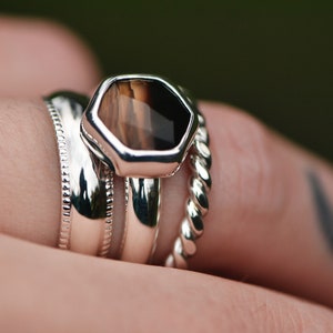 May include: A silver ring set with a hexagonal brown and black gemstone. The ring is stacked with two other silver bands.