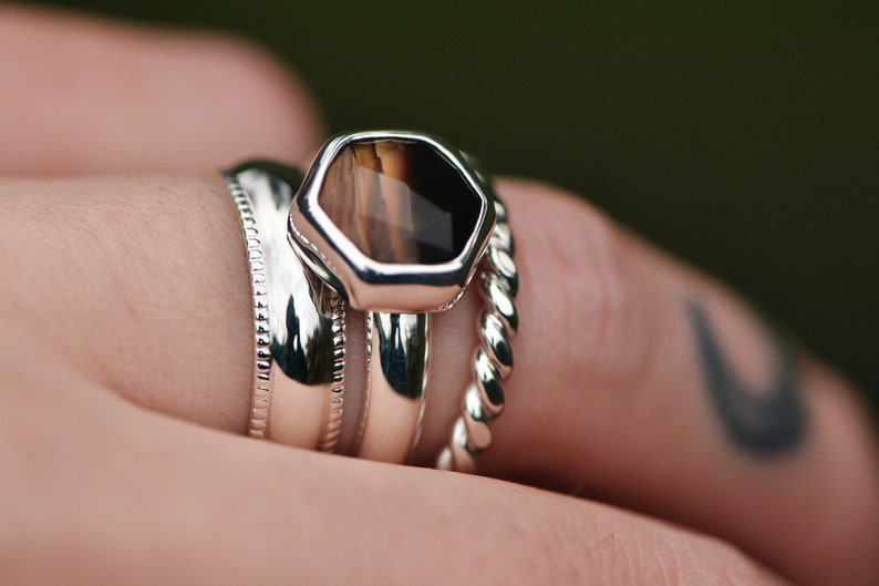 May include: A silver ring set with a hexagonal brown and black gemstone. The ring is stacked with two other silver bands.